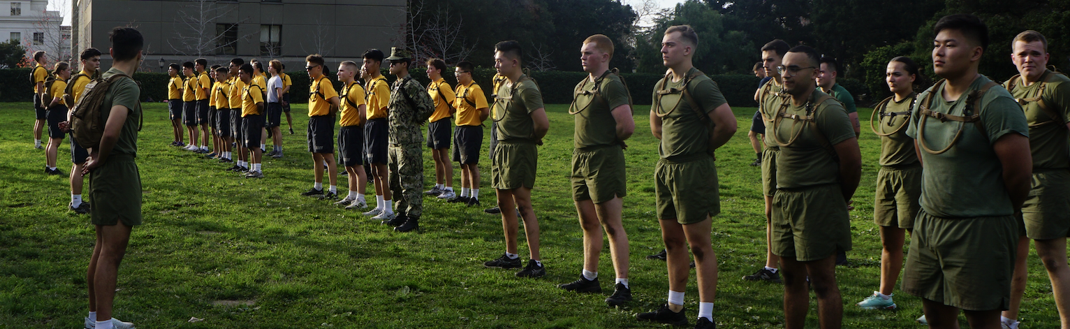 A large group of midshipmen on a grass field given instructions from the commanding officer