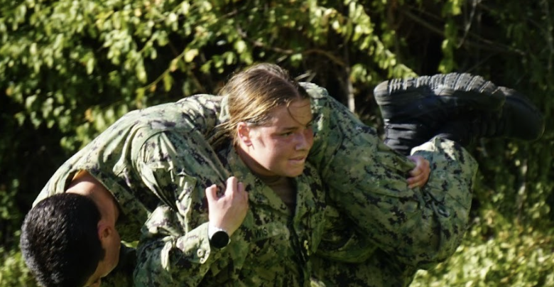 Female midshipmen in camouflage Navy working uniform training on the field carrying a male midshipmen over shoulder